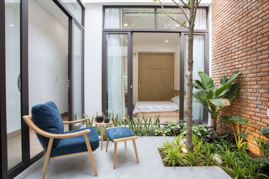 Taman indoor di samping rumah dengan desain minimalis, dihiasi tanaman hijau dan dinding semen ekspos.