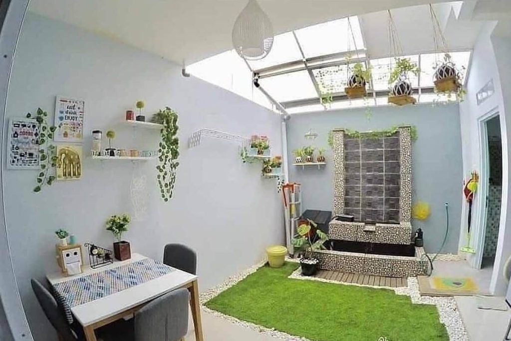 Taman indoor dengan rumput sintetis di belakang rumah, dikelilingi tanaman hias dan skylight yang memberikan cahaya alami.